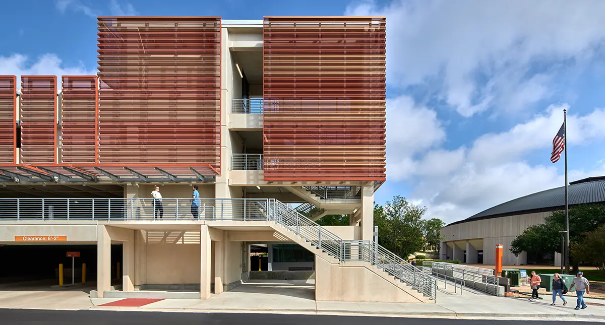 Modern building with red slatted panels and outdoor stairs on a clear day. People walk by, while an American flag waves nearby. Sky is partly cloudy.