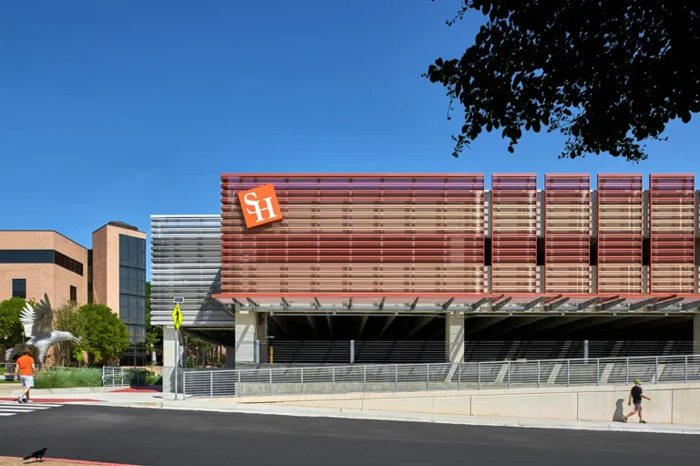 Modern building complex with SH logo, featuring red metal lattice facade. Bright blue sky, walkway in foreground, people walking, and greenery beside.