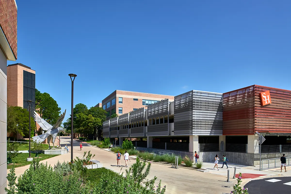 Campus scene with modern brick and metal buildings under a clear blue sky. Students walk along a path, and a large bird sculpture stands prominently.