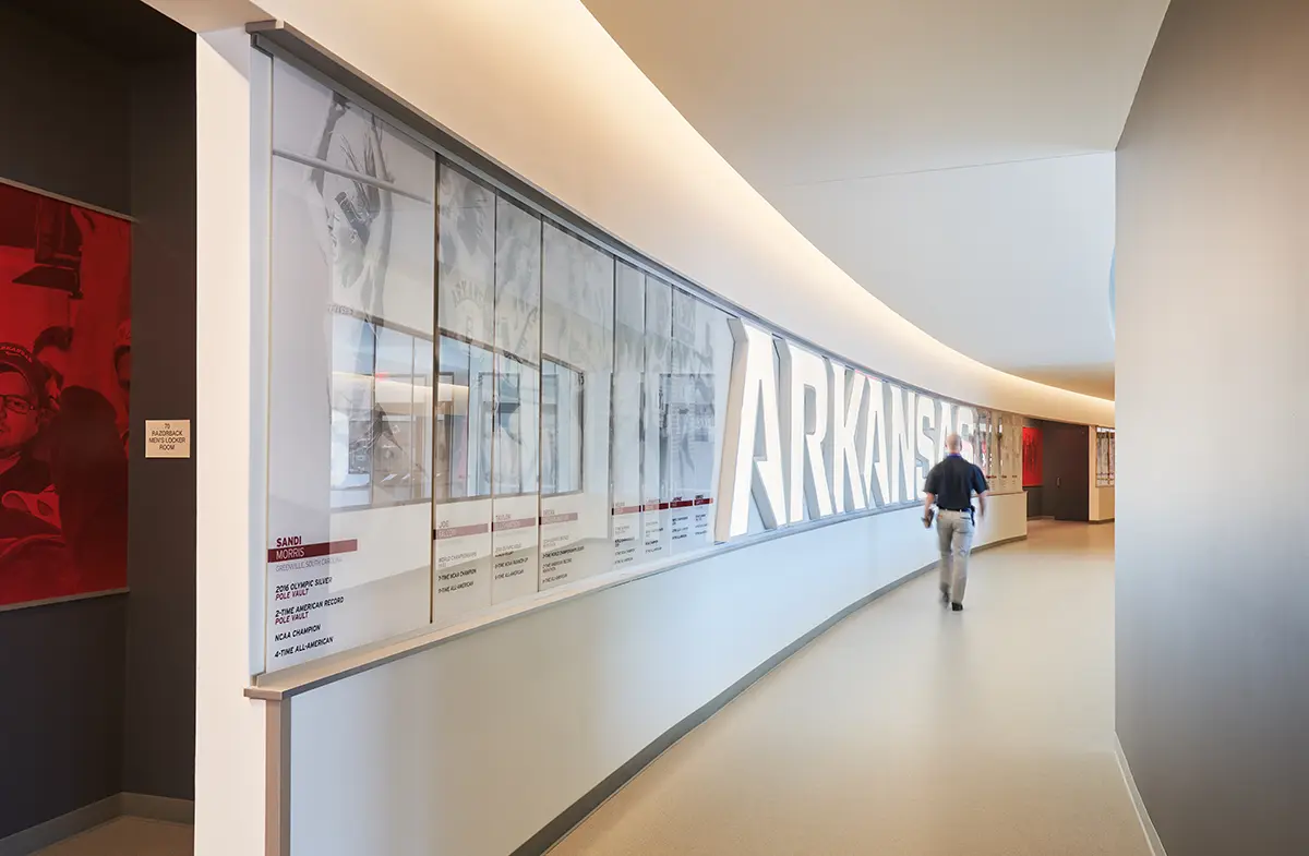 A man walks down a modern hallway with a large, backlit "ARKANSAS" sign on the wall. The design is sleek and bright, conveying a sense of innovation.