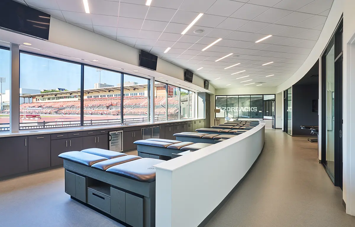 Spacious, modern athletic training room with treatment tables lined up along large windows overlooking a sports stadium, conveying a clean, professional atmosphere.