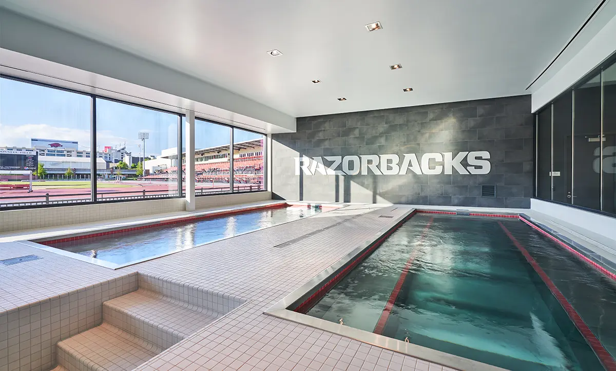 Indoor pool area with "RAZORBACKS" on a dark wall. Large windows reveal an outdoor stadium. The space feels modern and bright, suggesting a sports facility.
