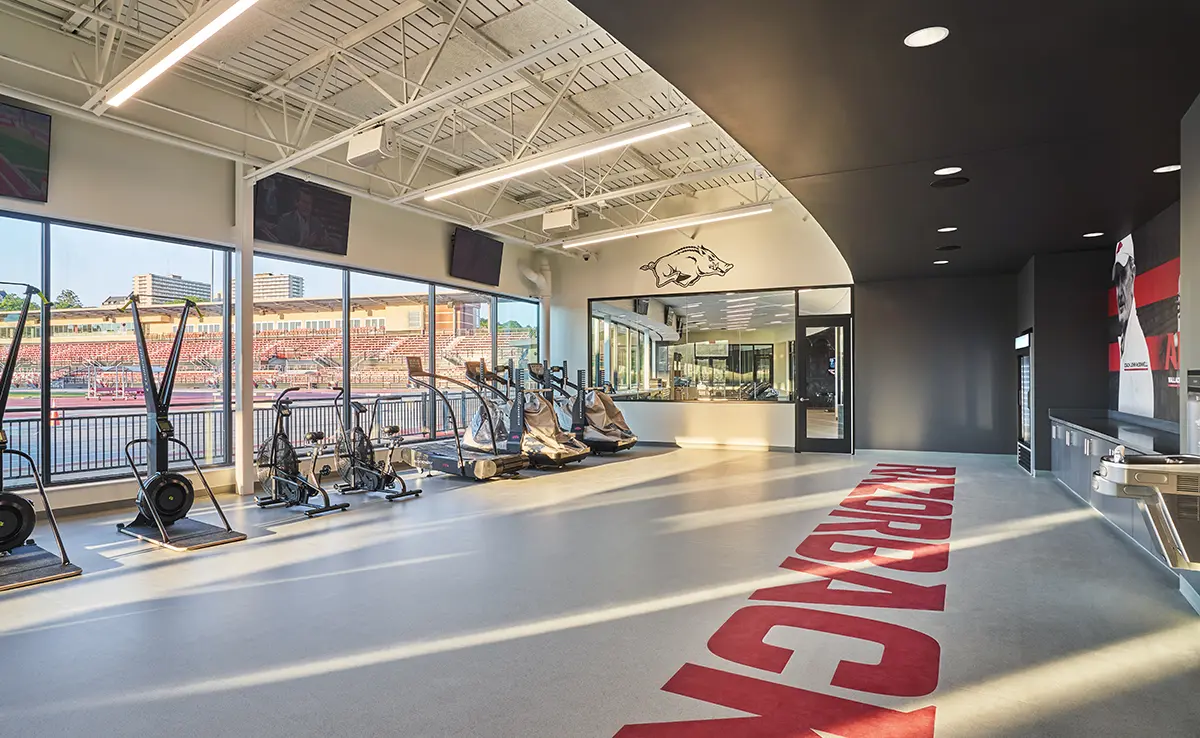 A bright, spacious gym with cardio equipment faces large windows overlooking a stadium. "RAZORBACK" is boldly written on the floor, creating an energetic atmosphere.