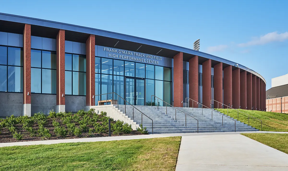 Curved brick building labeled "Frank O'Mara Track and Field High Performance Center." Glass entrance, wide steps, surrounded by greenery, clear blue sky.