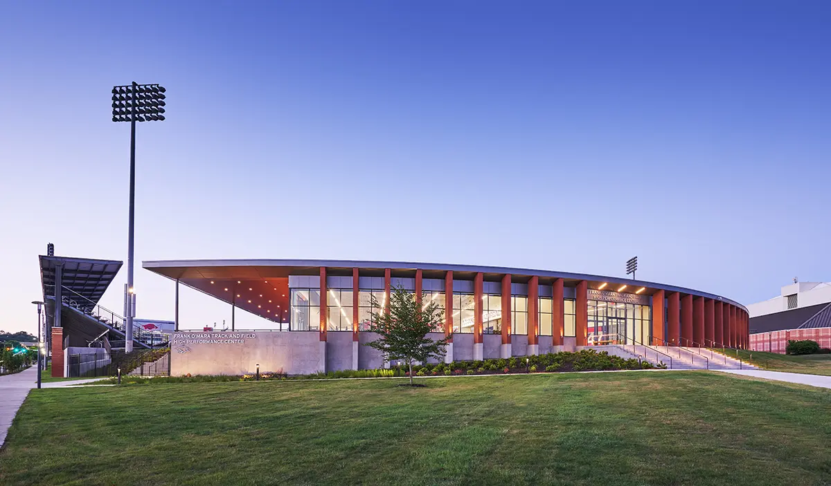 Sleek sports facility under a clear dusk sky, featuring modern architecture with tall glass windows and red pillars, surrounded by manicured green lawns.