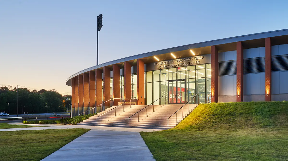 Modern building with tall glass entrance and illuminated steps, surrounded by green grass at dusk. Tall light poles and trees line the background.