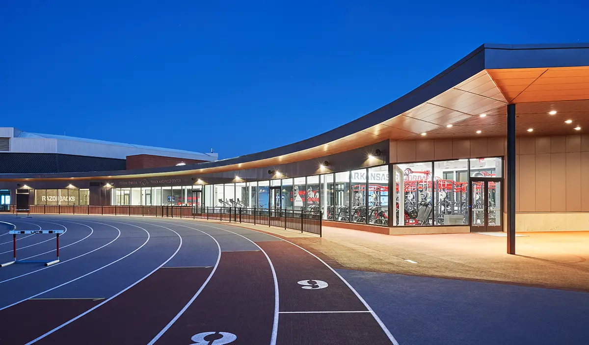 Curved building with large glass windows at dusk, revealing an indoor gym. Adjacent to an outdoor running track with numbered lanes. Calm and modern ambiance.