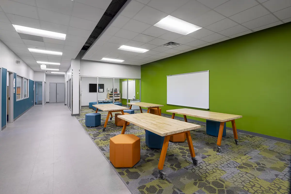 Modern classroom with wooden tables and colorful hexagonal stools on a patterned carpet. Green accent wall with a whiteboard creates a vibrant atmosphere.