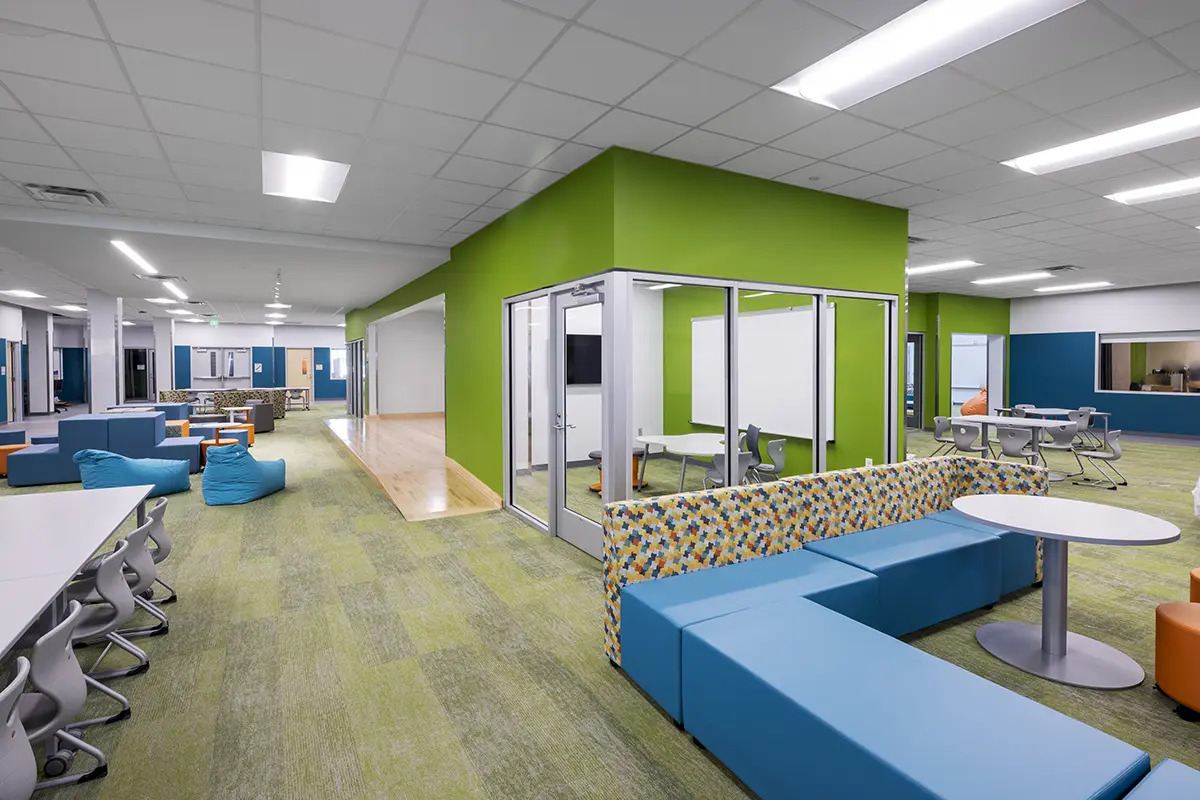 Modern classroom with a vibrant design. Features green walls, blue seating, a glass-walled meeting room, tables, and a casual, collaborative atmosphere.