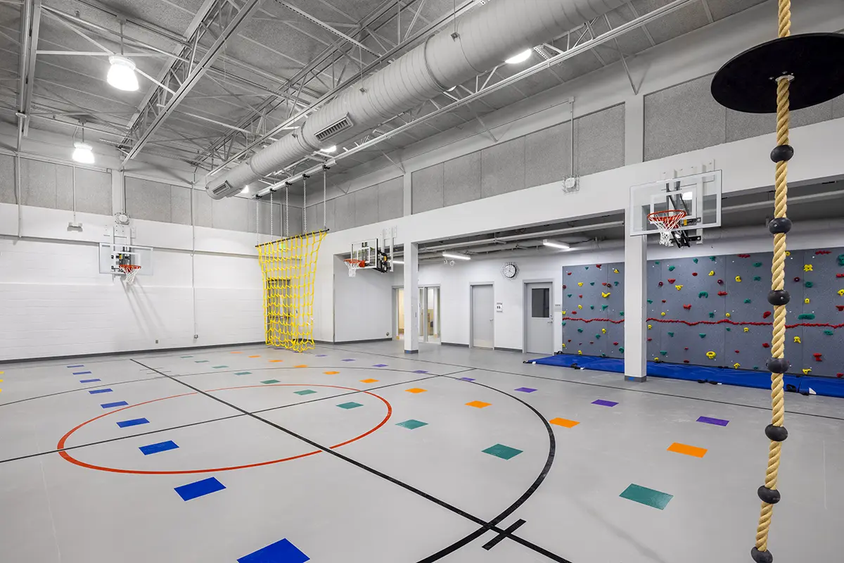 A bright gymnasium featuring a climbing wall, basketball hoops, a rope with a disc, and colorful floor markings. The atmosphere is energetic and inviting.