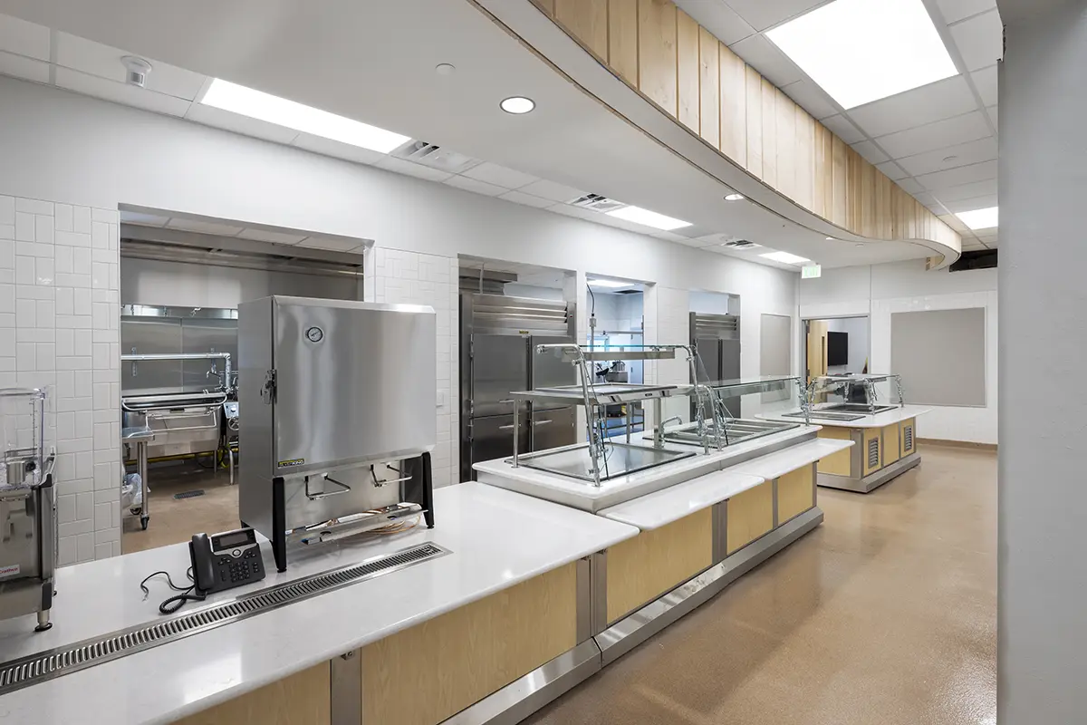 A modern, empty cafeteria serving area with stainless steel appliances, glass display cases, and wooden accents under bright fluorescent lights. Clean and orderly.