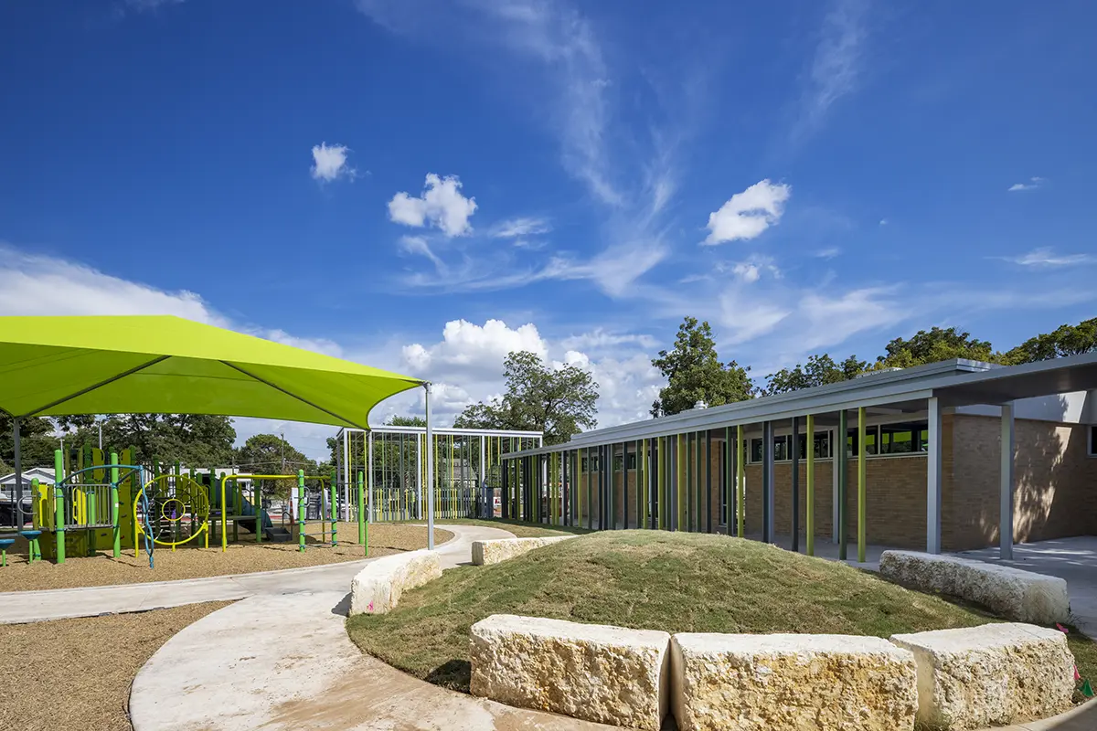 Modern playground with a vibrant green shade structure beside a school building. Sunny weather, blue skies with clouds, and a welcoming atmosphere.