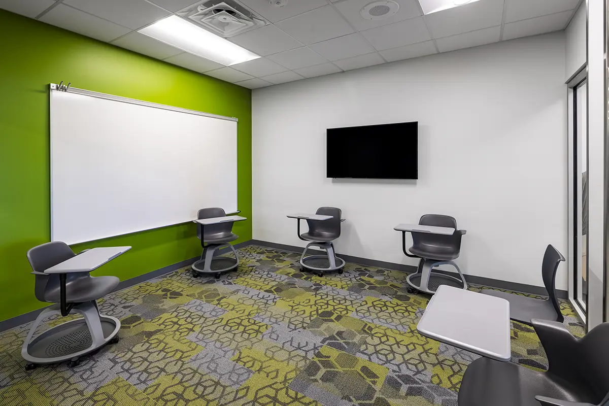 Small modern classroom with green and white walls, four swivel chairs with attached desks, a wall-mounted screen, and a colorful patterned carpet.