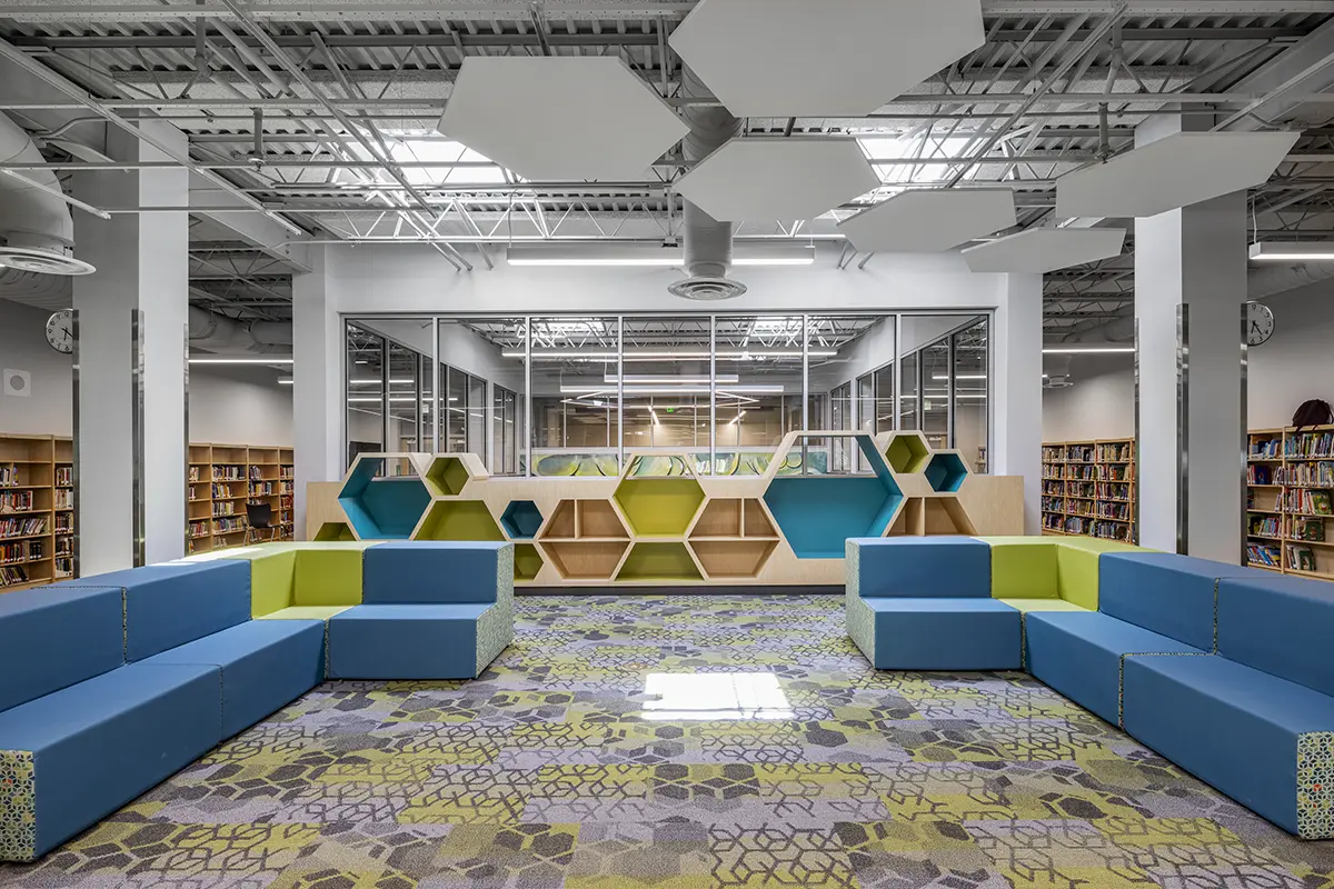 Modern library interior with colorful geometric seating in blue and green. Hexagonal shelves line the wall, and patterned carpet covers the floor. Bright and inviting atmosphere.