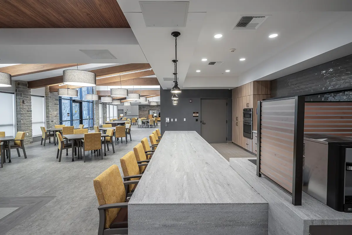 Spacious dining area with a long stone counter in the foreground, yellow chairs, wooden tables, large ceiling lights, and natural light from windows.