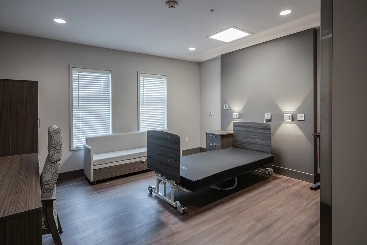 A modern hospital room with a minimalist design, featuring a raised hospital bed, wooden floors, soft lighting, two windows with blinds, and a patterned chair.