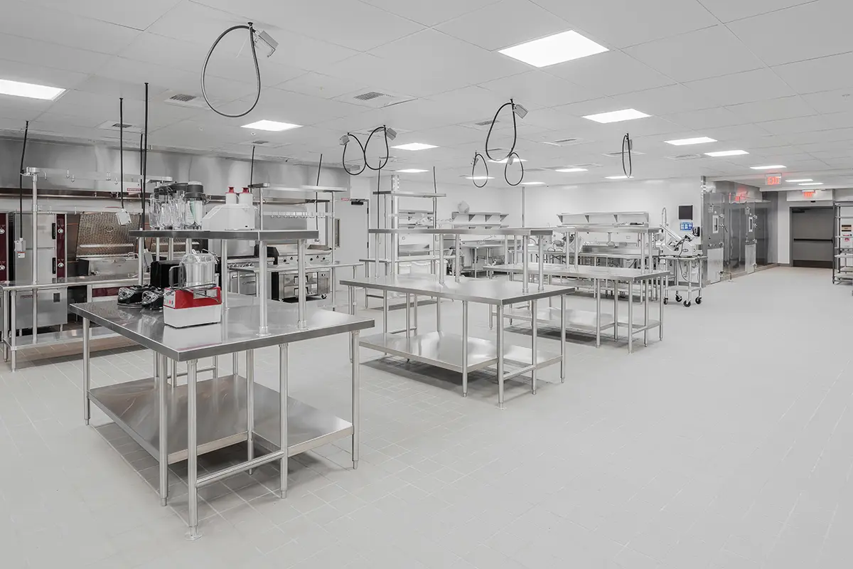 Spacious industrial kitchen with stainless steel worktables, ovens, and hanging hoses. Bright ceiling lights create a clean, organized atmosphere.