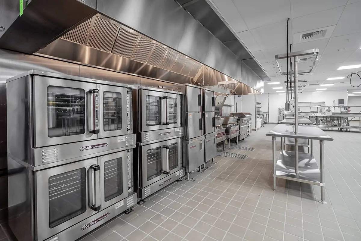 Spacious commercial kitchen with stainless steel ovens, grills, and tables. Clean, organized, and modern with a focus on efficiency and hygiene.