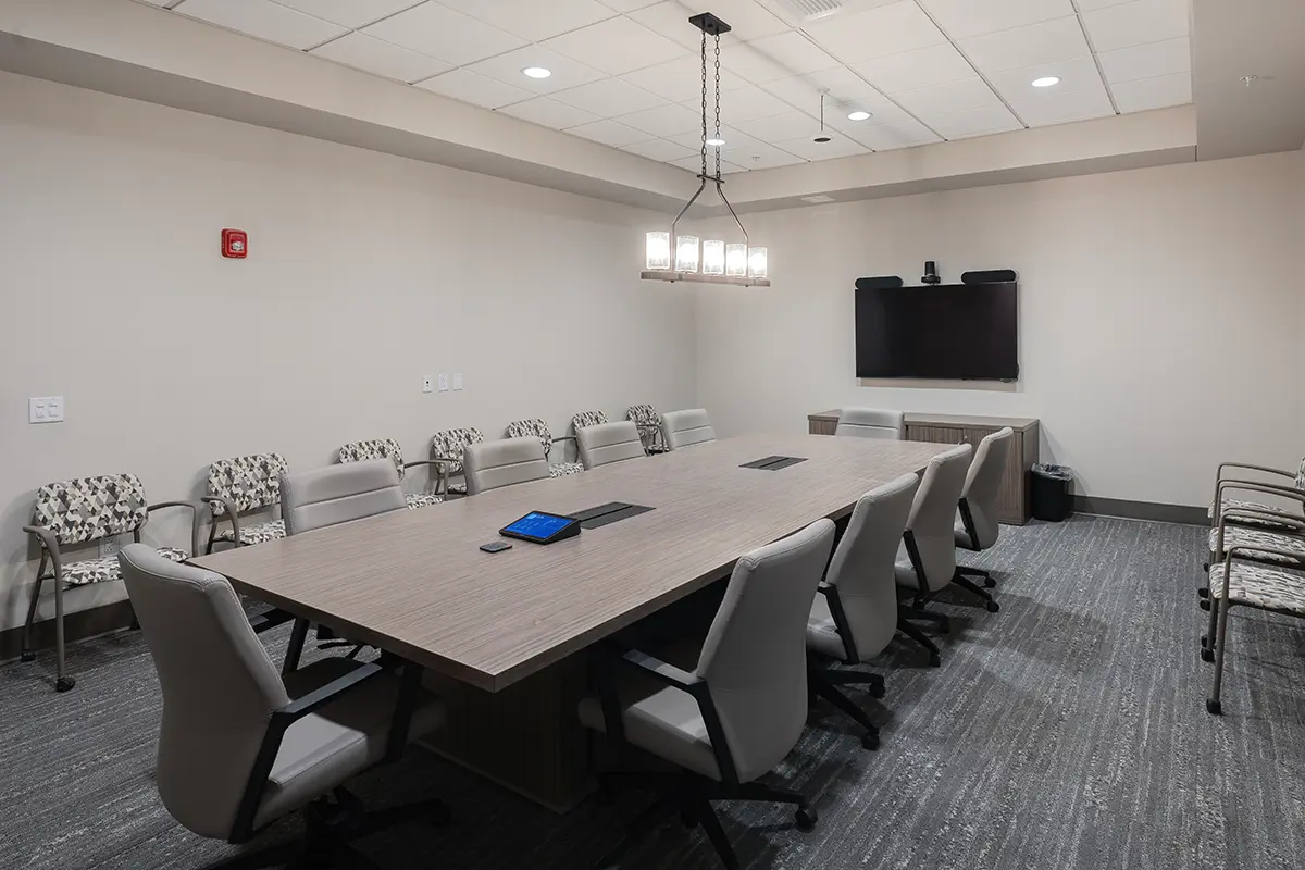 A modern conference room with a long wooden table surrounded by gray chairs. A TV is mounted on the wall, and a chandelier provides soft lighting. The room is neat and professional.