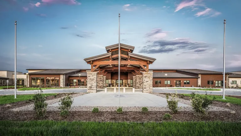 A modern building with a stone and wood entrance under a clear blue sky with pink clouds. Three flagpoles and landscaped greenery are in the foreground.