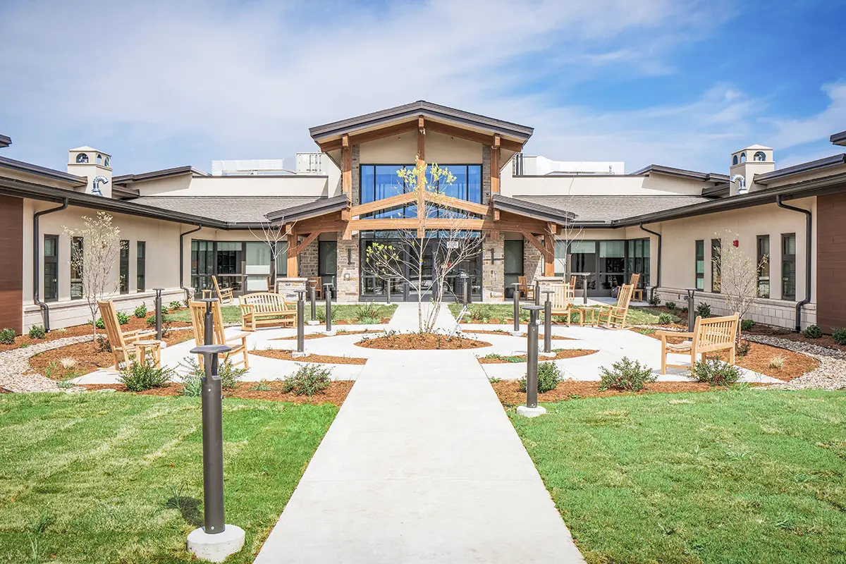 Modern building with a peaked wooden entry, surrounded by landscaped gardens. Benches line the walkway, creating a welcoming and serene atmosphere.