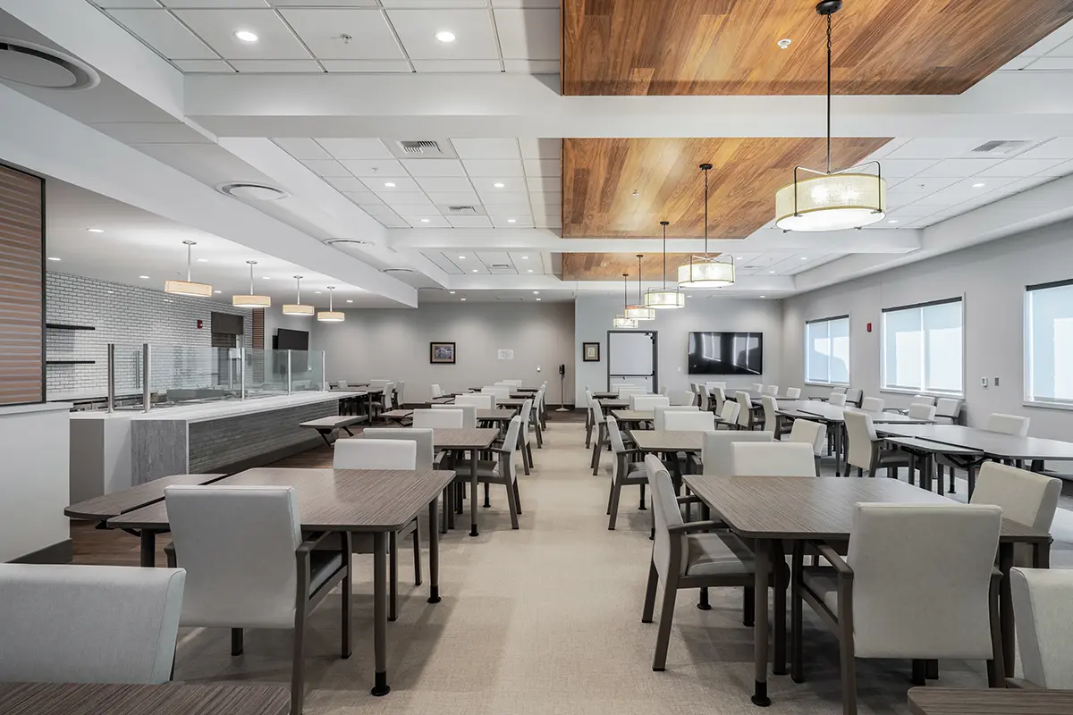 Modern dining area with neatly arranged wooden tables and cushioned chairs under pendant lights. A calm, spacious atmosphere with large windows and neutral colors.