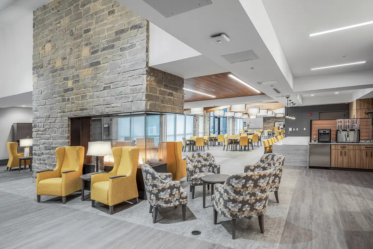 Spacious modern lounge with stone fireplace, yellow armchairs, floral-patterned chairs, and dining tables in the background. Bright, inviting atmosphere.