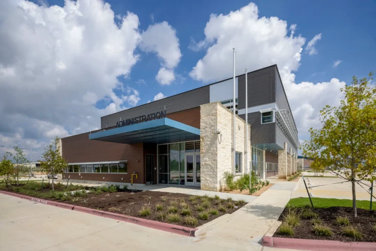 Modern two-story building labeled "Administration" with large stone and metal facade, landscaped greenery, under a blue sky with scattered clouds.