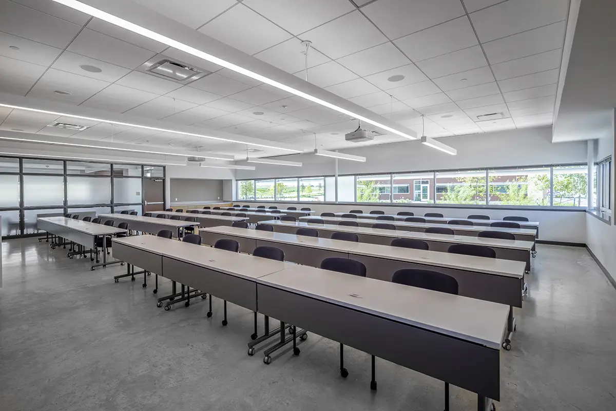 Spacious, bright classroom with long rows of empty desks and chairs. Large windows let in natural light, creating an open and inviting atmosphere.