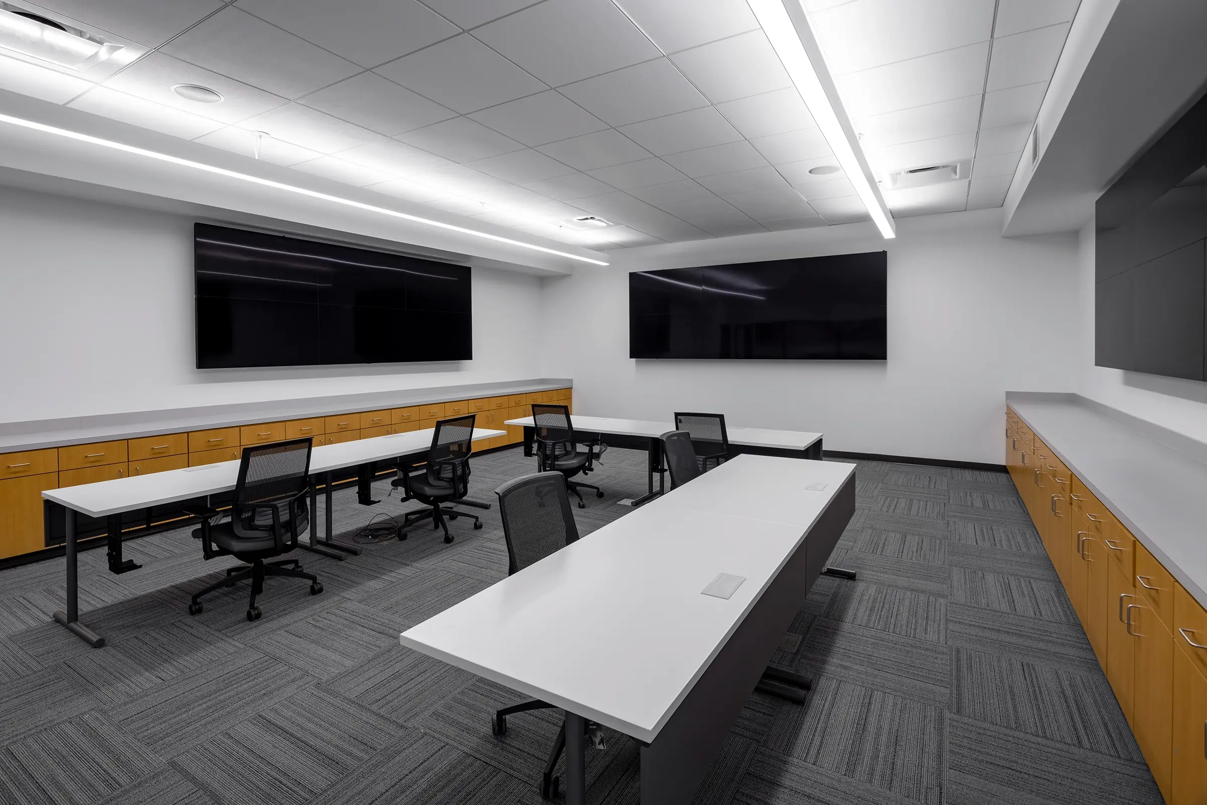 A modern conference room with sleek white tables, black mesh chairs, and two large wall-mounted screens. Cabinets line walls under low, bright lighting.