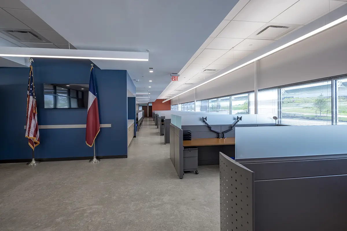 Modern office interior with sleek cubicles under bright lighting. U.S. and Texas flags displayed by a blue wall. Large windows offer natural light.