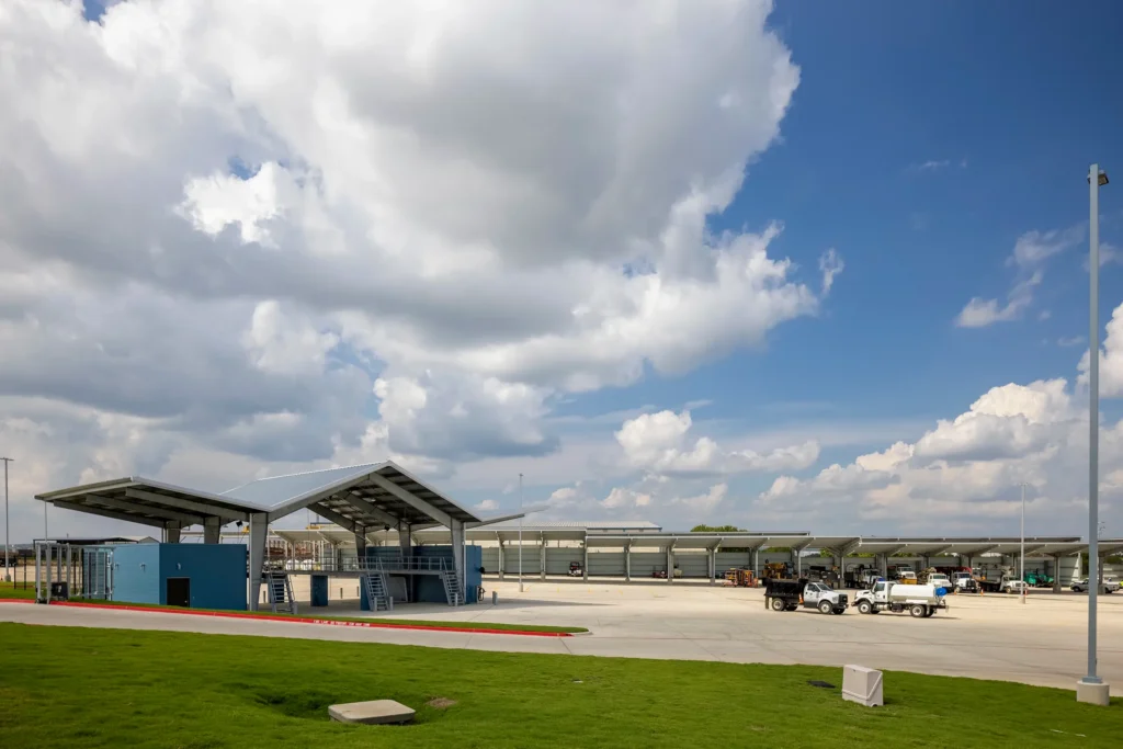 A modern industrial facility under a partly cloudy sky. The scene shows a large paved area with some parked trucks and a small building with a sloped roof.
