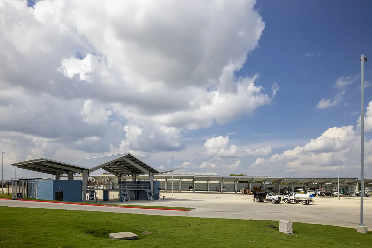 Large open parking lot under a bright blue sky with fluffy clouds, featuring a small blue utility building with a metal roof in the foreground.