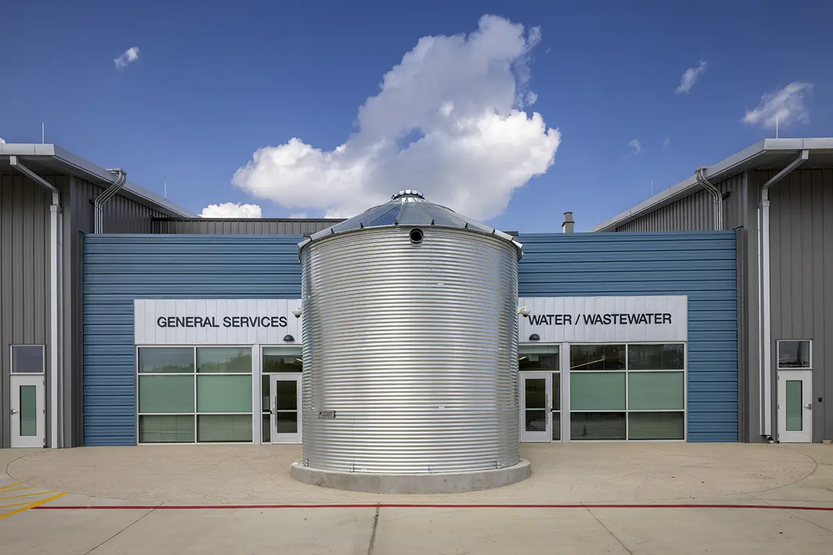 Modern industrial building with blue and gray facade, featuring a large silver cylindrical tank in front. The signs read "General Services" and "Water/Wastewater." The scene is bright under a blue sky with fluffy clouds.