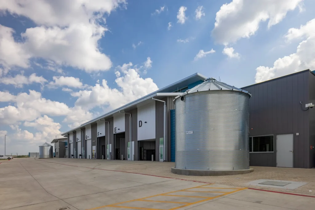 Industrial warehouse with a row of open bays labeled D and E under a bright sky. A large metal silo is prominently in the foreground.