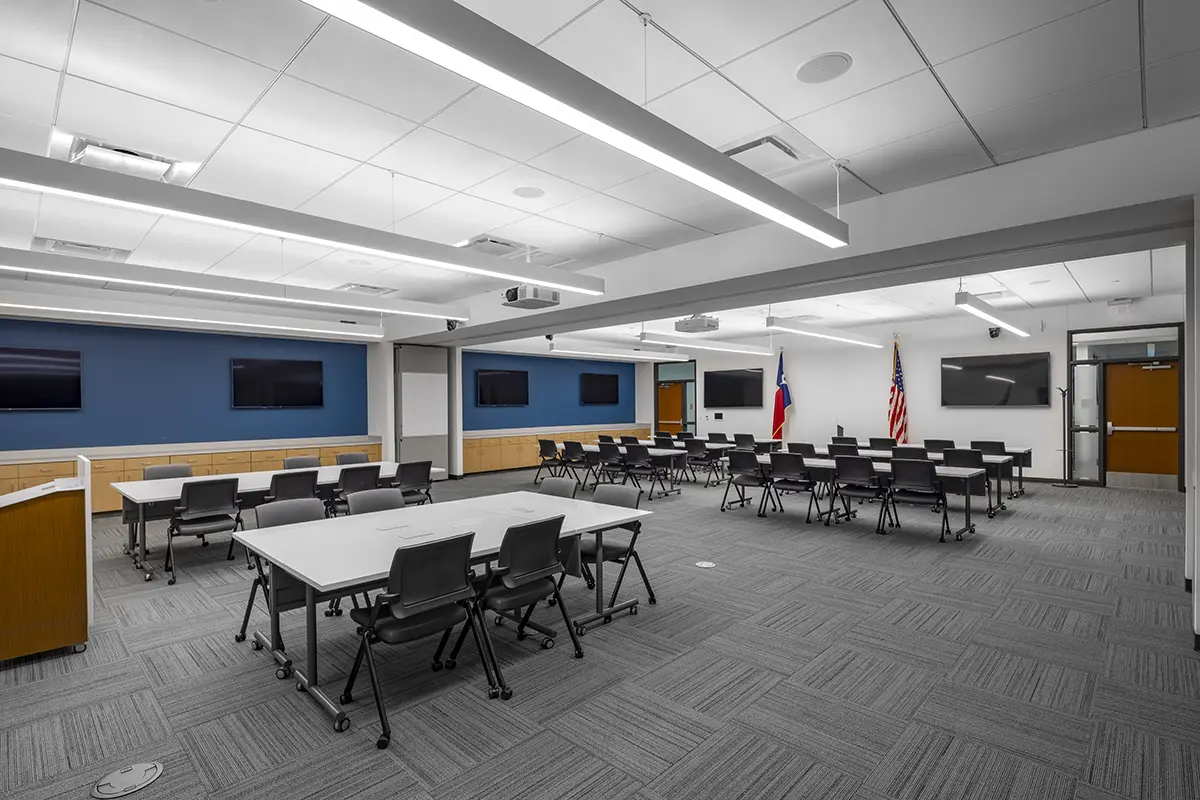 A modern conference room with gray carpet, white walls, and blue accent wall featuring multiple TVs. Tables and chairs are arranged for meetings. American and Texas flags stand in the corner. Bright overhead lighting creates a professional atmosphere.