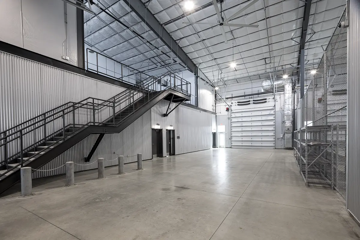 Spacious industrial facility interior with high ceilings, a staircase, large metal doors, and bright lighting. The atmosphere is clean and utilitarian.