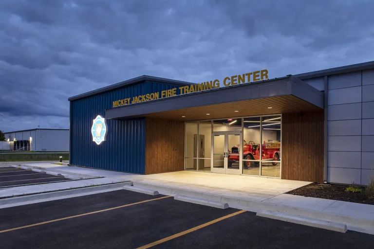 Modern fire training center with illuminated logo and vintage fire truck seen through glass doors. The exterior is sleek, with a calm evening sky.