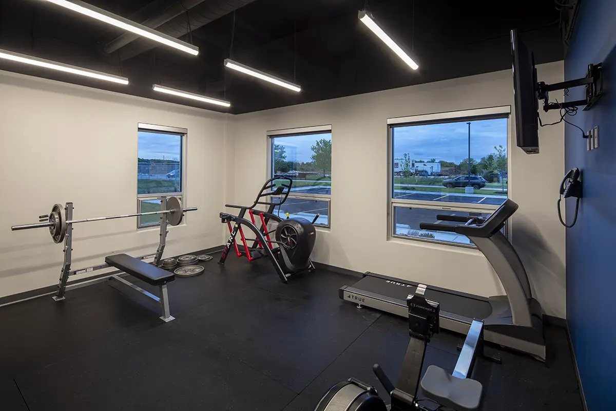 A modern gym room with large windows and natural light. Equipment includes a bench press, exercise bike, treadmill, and rowing machine. The ambiance is clean and inviting.