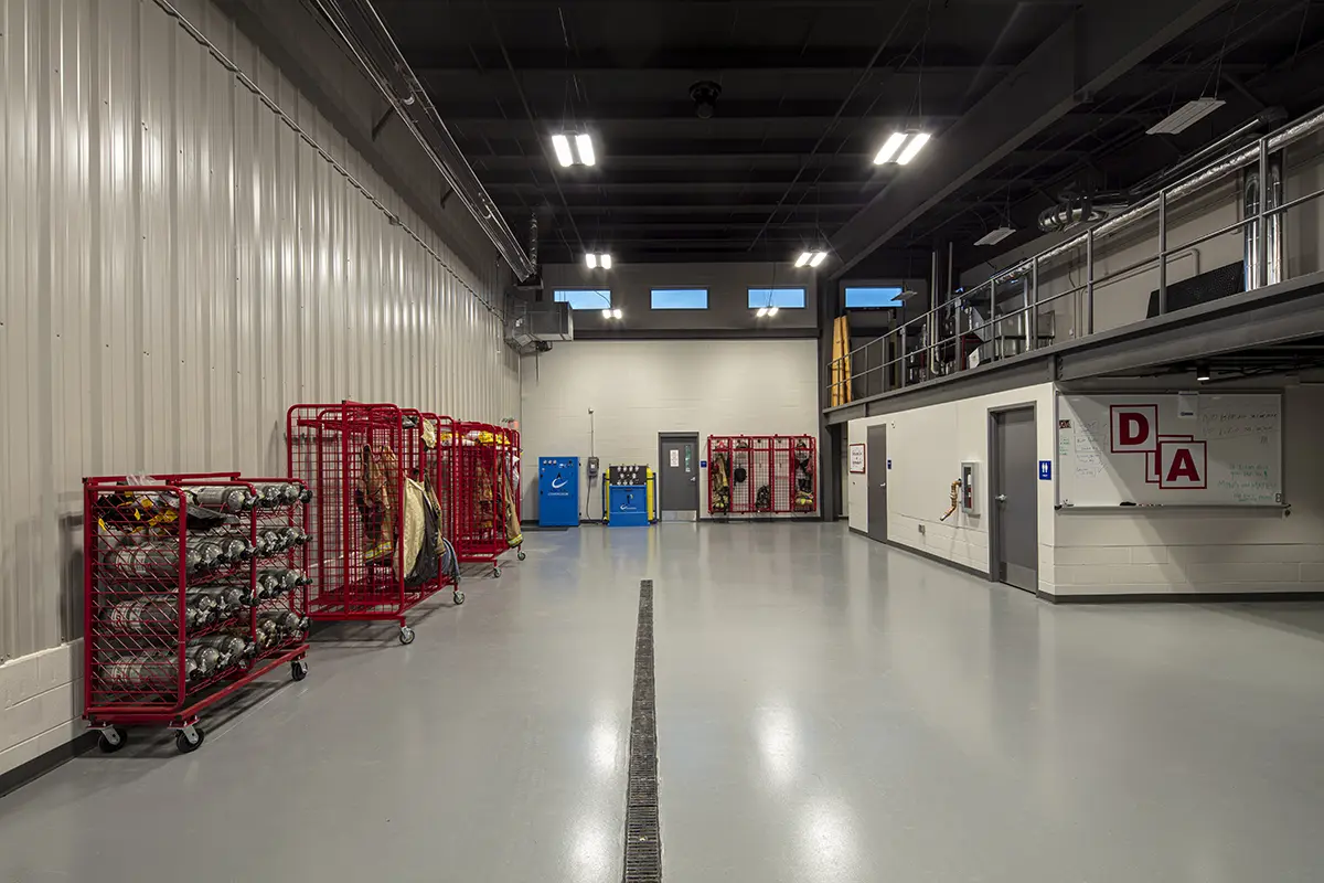 A spacious industrial room with gray walls and floor, featuring red storage racks with equipment on the left. Bright overhead lights create a well-lit, organized atmosphere.
