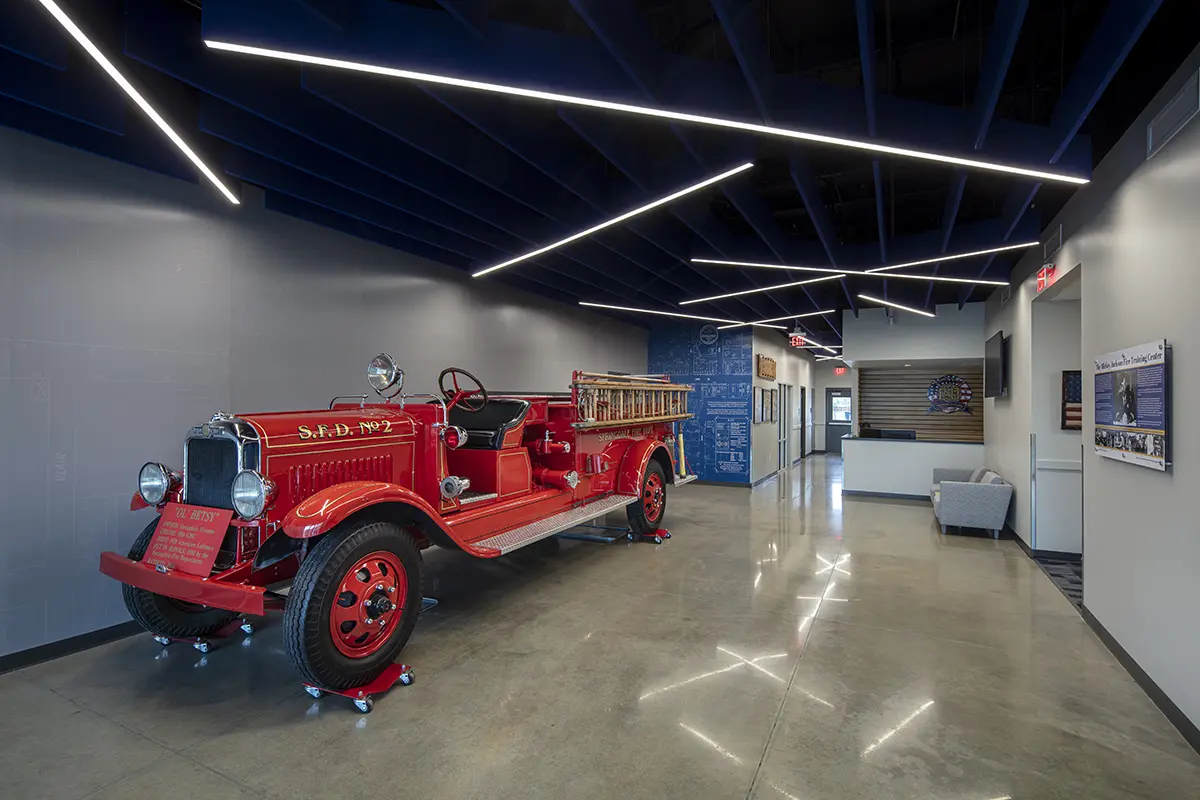 A vintage red fire truck is displayed in a modern lobby with sleek gray walls and dynamic overhead lighting, creating a blend of history and innovation.