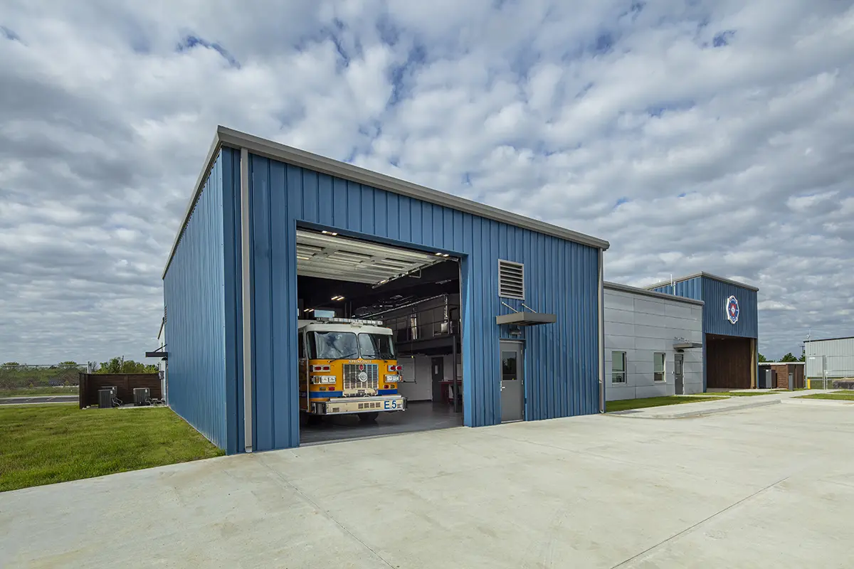 A bright fire truck is parked in a blue metal garage under a partly cloudy sky. The scene is calm, with grassy areas surrounding the station.