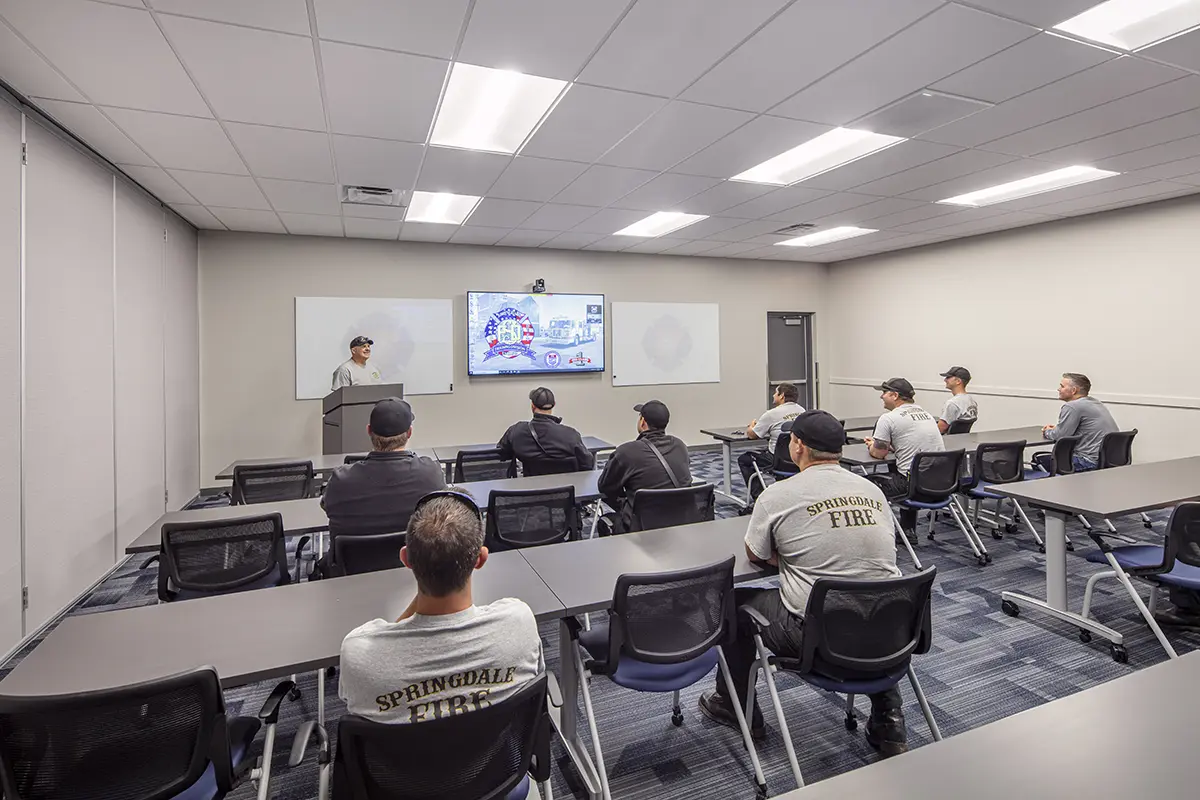 A group of firefighters in uniform attend a training session in a modern classroom. They face a large screen with a presenter, conveying focus and learning.