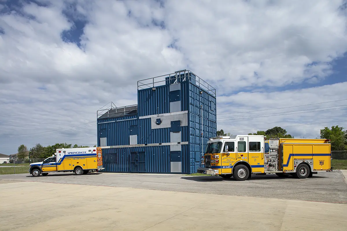 Fire training facility with blue metal structure, yellow fire truck, and ambulance parked in front. Overcast sky adds a sense of readiness and purpose.