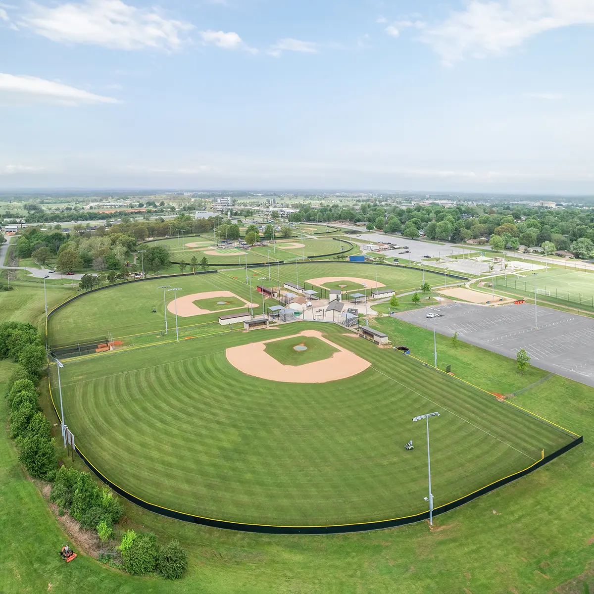Aerial view of multiple baseball fields surrounded by lush green grass. The fields have well-maintained, symmetrical layouts under a partly cloudy sky.