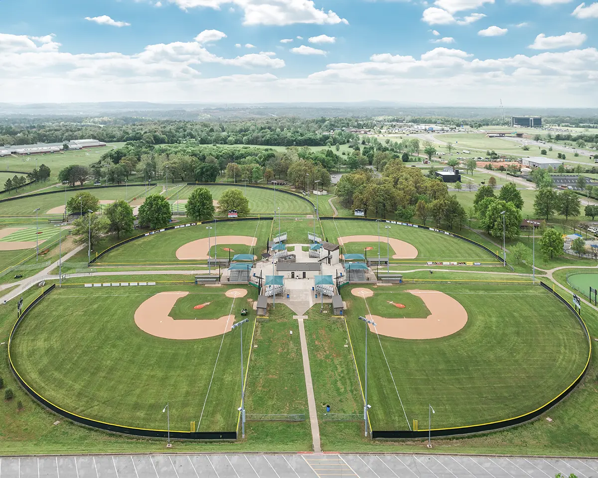 Aerial view of a baseball complex with four symmetrical fields, lush green grass, and surrounding trees under a bright blue sky with scattered clouds.