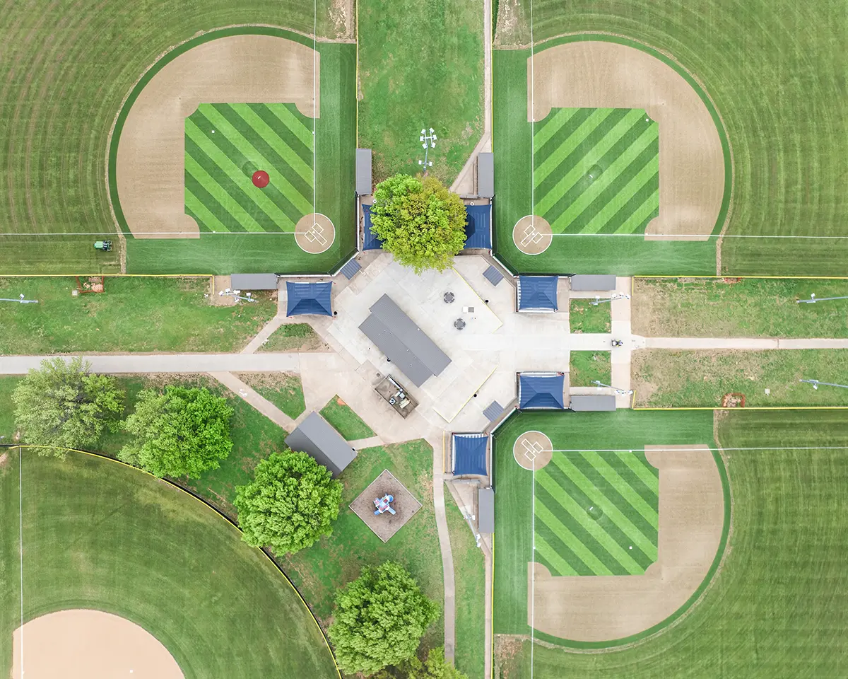 Aerial view of a symmetrical sports complex with four baseball fields, lush green grass, dirt infields, central seating area, and surrounding trees.