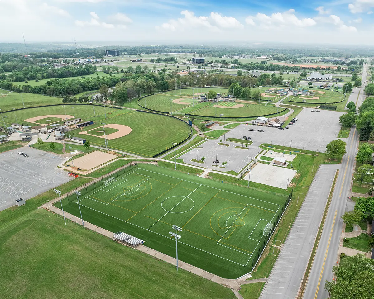 Aerial view of a sports complex featuring a green soccer field and multiple baseball diamonds. The area is surrounded by parking lots and trees.