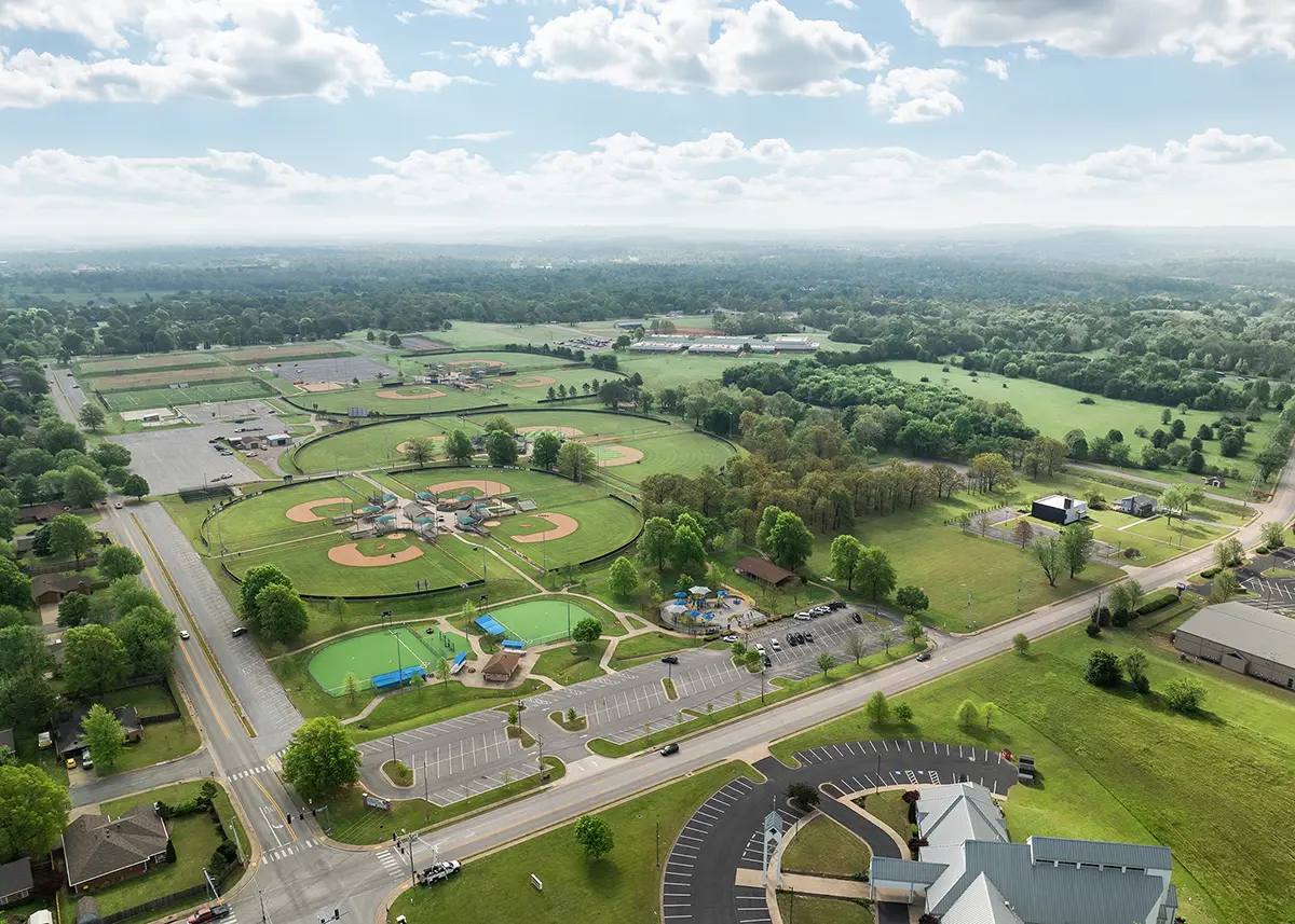 Aerial view of a park with multiple baseball fields, adjacent roads, and parking. Lush greenery and a distant horizon under a partly cloudy sky.