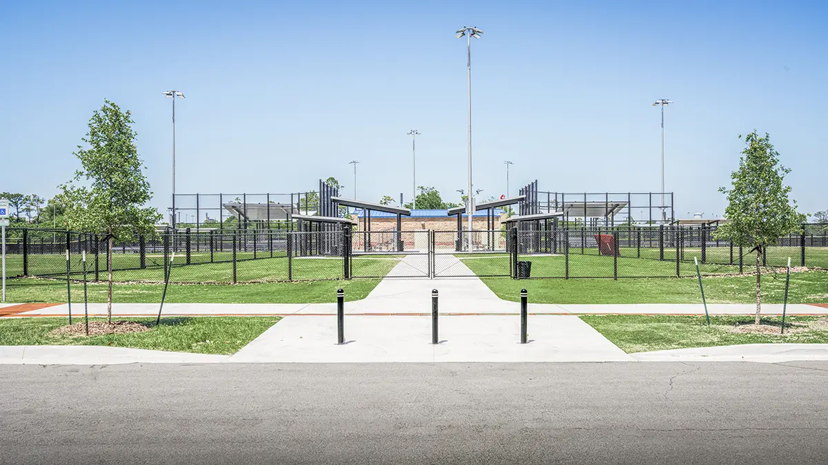 Wide view of a baseball field entrance, surrounded by green grass, trees, and tall floodlights under a clear blue sky. The setting appears calm and inviting.